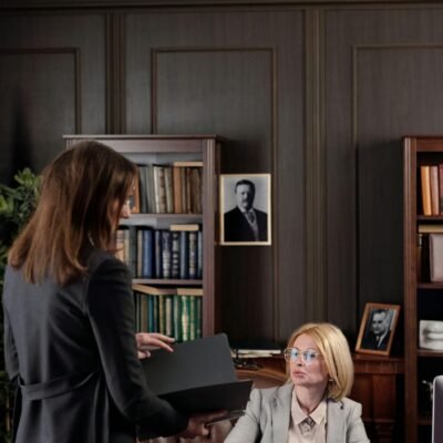 Two businesswomen at a meeting in a book-filled office. Professional and focused setting.